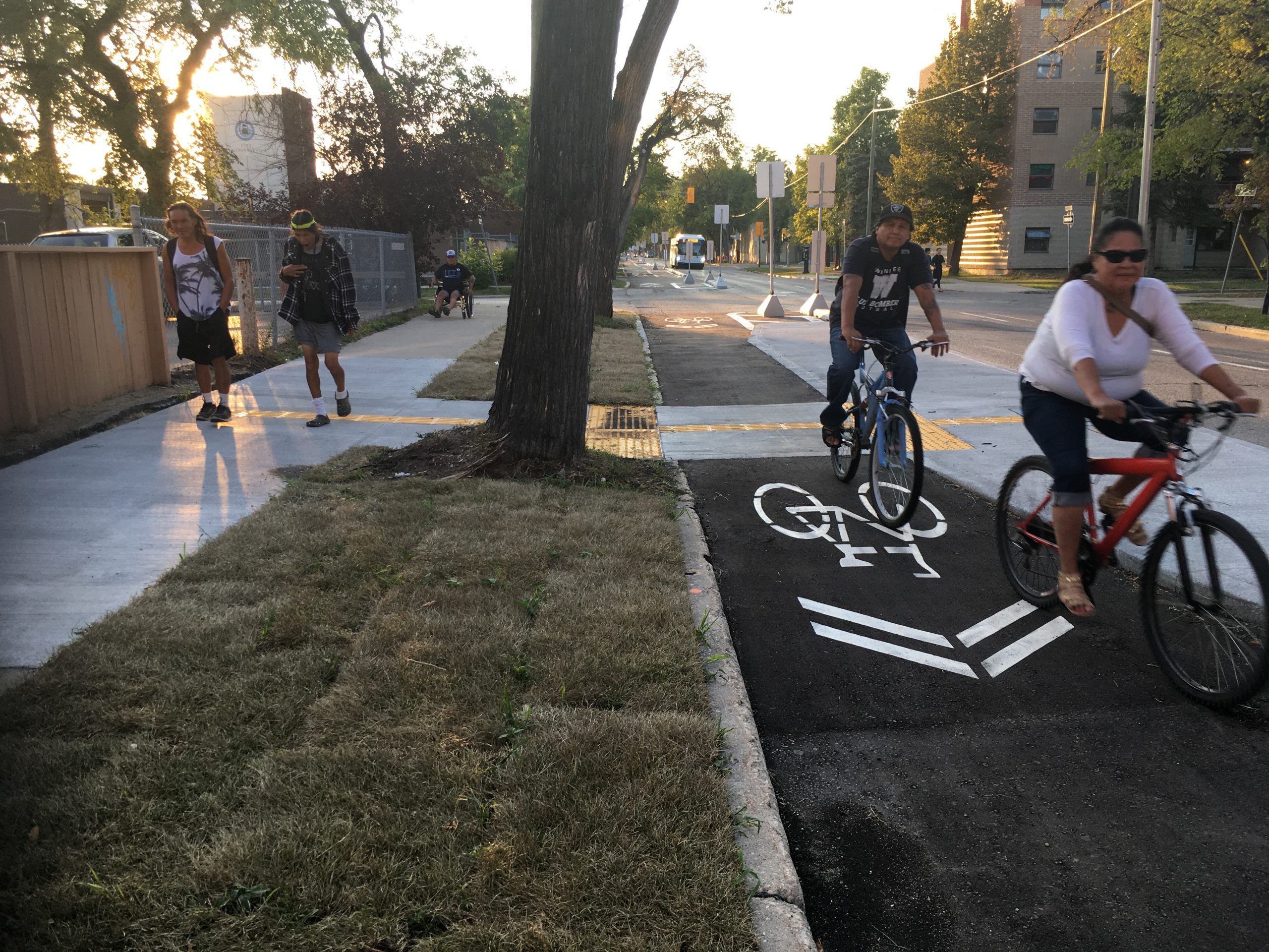 Photo of new sparated bike lane (Fall 2018) showing a couple riding, beside a sidewalk showing a couple waking and one person in a wheelchair.